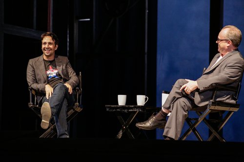 Two men sit cross-legged and comfortably laughing in director chairs on stage