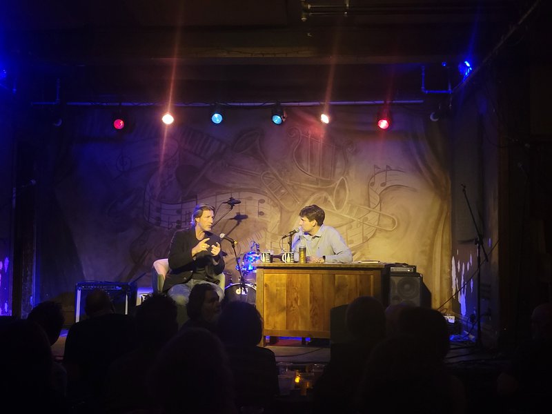 Two white men sit chatting on a brightly colored lit stage at a dark bar. The man to the left has casually slicked back brown hair and wears a black coat and jeans, the man to the right sits behind a desk and wears a blue button down.