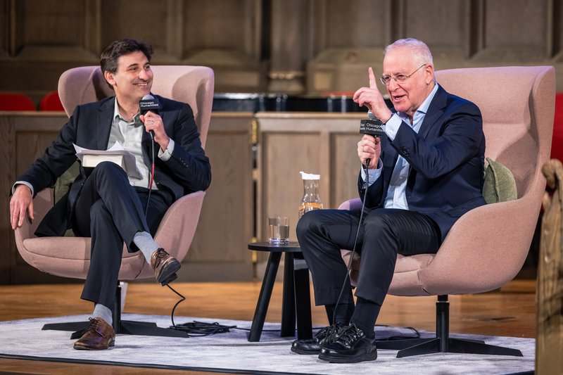 Ron Chernow and Mark Bazer sit on stage speaking into hand-held microphones with a small table between them. Seated on the left, Mark Bazer has dark brown hair, grey shirt, navy blue suit, and brown shoes. While gesturing with his left hand index finger pointed up, Ron Chernow has grey hair and glasses. He is wearing black shoes with a blue shirt, pants, and sportscoat.