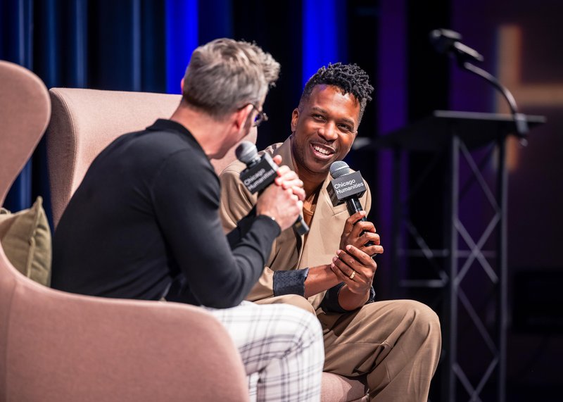 A photo of a Black man smiling, and looking at the man interviewing him intently. He holds a microphone that reads “Chicago Humanities” and wears a chic flowy light brown suit. The back of the head of a white man with silver hair is seen listening. He wears a black shirt and white and gray plaid slacks, and also holds a microphone.