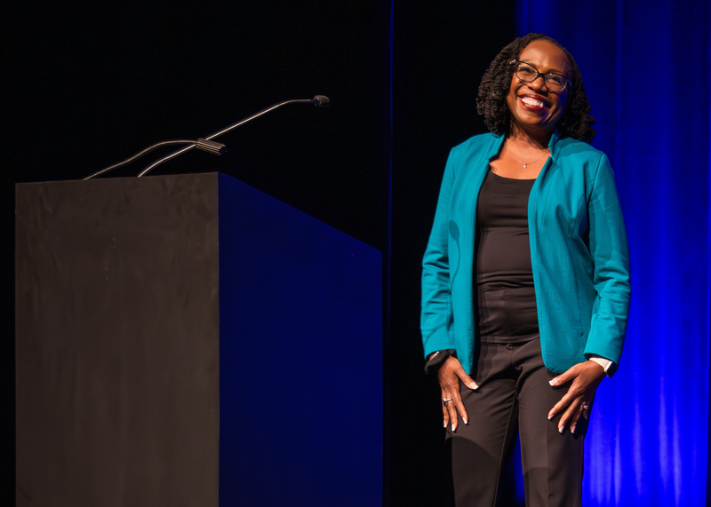 A beaming Black woman stands on stage, just to the side of a podium. She has a big smile with red lipstick, with bobbed hair in braided curled twists. She wears a bright teal blazer over black pants and top and oval cat eye glasses.