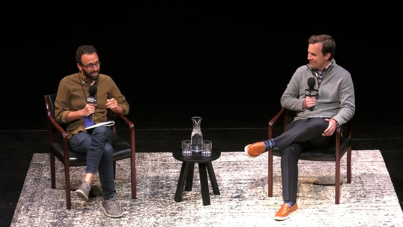 Two men sit in studious wooden chairs on a stage with a nice rug, with legs crossed and holding microphones. The man on the left has glasses and a beard, and gestures to make a point. The man on the right wears a gray zip up and smiles.