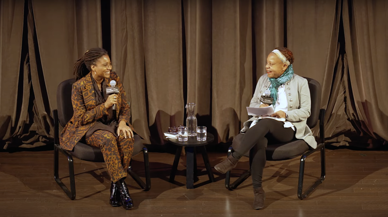 Two Black women sit on a wooden stage with a thick, shiny brown curtain behind them. The woman on the right has dreadlocks and wears a brown patterned suit and black shiny boots, talking and smiling. The woman on the right wears a green scarf and gray jacket with big buttons, and smiles, holding papers.